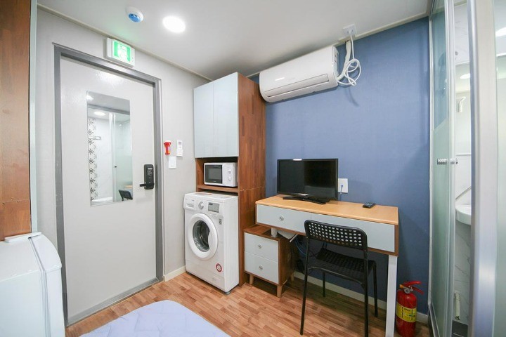 部屋の内部が描かれています。左側には洗濯機があり、その隣には電子レンジが置かれたキャビネットがあります。右側にはデスクと椅子があり、テレビがデスクの上にあります。空調設備が天井に取り付けられており、ドアには小さな窓があります。明るい照明があり、清潔感のある居住空間です。