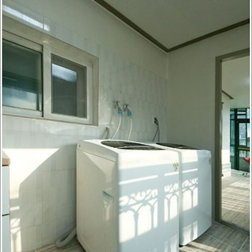 白い洗濯機が部屋の中央に置かれている様子が写っています。部屋の壁には窓があり、窓からの光が部屋に差し込んでいます。床は木製のフロアで、洗濯機の周りには白い壁が見られます。部屋の雰囲気は明るく清潔な感じです。