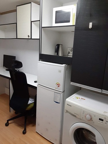 シンプルなデザインの小さな部屋。左側には黒いオフィスチェアとデスク、デスクの上にはモニターがあります。右側には白い冷蔵庫と洗濯機、洗濯機の隣には電子レンジがあります。壁には棚があり、収納スペースが確保されています。