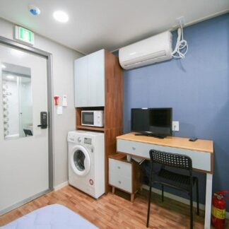 部屋の内部が描かれています。左側には洗濯機があり、その隣には電子レンジが置かれたキャビネットがあります。右側にはデスクと椅子があり、テレビがデスクの上にあります。空調設備が天井に取り付けられており、ドアには小さな窓があります。明るい照明があり、清潔感のある居住空間です。