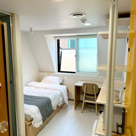 天窓から光が差し込む明るいワンルームの室内写真です。白いベッドにはグレーのブランケットがかけられ、清潔感があります。窓際には机と椅子が置かれ、学習や作業スペースとして利用できそうです。部屋の片側には収納棚が設置され、収納力も確保されています。全体として、シンプルで快適な生活空間が感じられます。