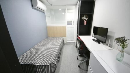 白い壁と木製の家具がある、明るい居室の写真です。部屋には大きなベッド、コンピューターデスク、本棚があり、居心地の良い雰囲気が感じられます。オフィスや寝室として使えそうな空間ですね。