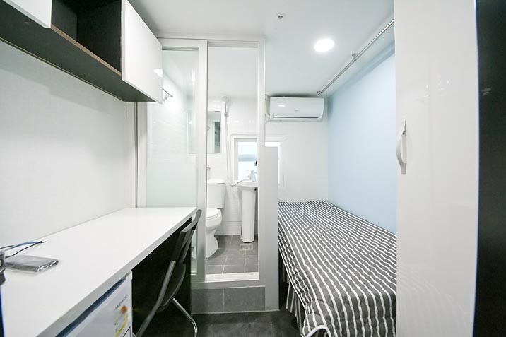 白い壁の部屋には、ストライプ模様の布団が掛けられたベッドと、机が配置されている。机の上には小物があり、向かい側にはガラスで仕切られたトイレが見える。部屋の奥にはエアコンが取り付けられている。