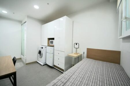 白い部屋に洗濯機と冷蔵庫が置かれた設備の整ったリビングルームの画像ですね。ベッドの上には縦縞のシーツが敷かれており、部屋全体が清潔で整理されています。