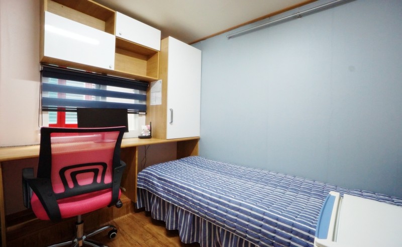 部屋の内部が映った画像です。シンプルなデザインのベッドがあり、ストライプ模様の布団が掛けられています。ベッドの隣には木製のデスクがあり、その上には黒い椅子が置かれています。窓にはブラインドがあり、壁は淡い青色です。壁には収納キャビネットもあります。全体的に落ち着いた雰囲気のある空間です。