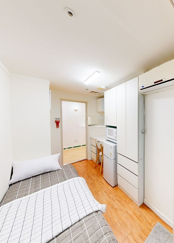 部屋の内部が写っており、左側には白い壁に掛けられた赤い非常ベルが見えます。中央には二つのベッドが並んでおり、一方には白い枕とグレーのブランケットが置かれています。右側には冷蔵庫、電子レンジ、そして収納キャビネットが並んでいます。奥にはドアがあり、その向こうには明るい空間が見えます。全体的に清潔感のあるシンプルなデザインの部屋です。