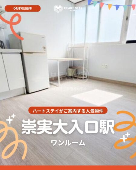 明るい日差しが差し込む、白を基調としたワンルームの室内。左側にキッチンと冷蔵庫、奥の窓際に小さなデスクが置かれています。