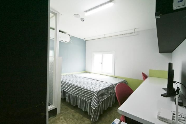 ストライプ模様の布団がかけられたベッドがあり、左側にはシャワールームが見える。部屋の奥には小さな窓があり、自然光が入る。デスクと赤い椅子が配置され、シンプルで明るい内装の部屋。