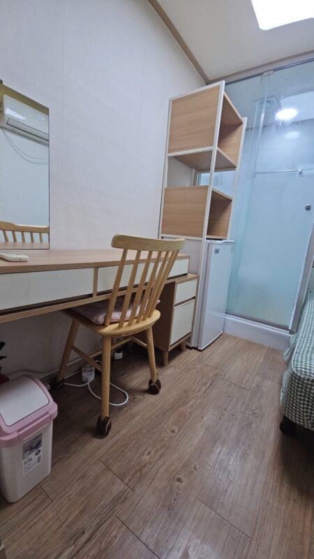 部屋の中には、机と椅子が置かれています。机の上には、コンピューターやその他の物品が置かれています。部屋の奥には、棚やタンスなどの収納家具が設置されています。部屋全体が明るく、清潔な印象を受けます。