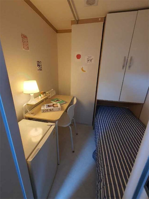 部屋の一角が映った画像です。左側には小さな机と椅子があり、デスクランプが点灯しています。机の上には本や文房具が置かれています。右側にはストライプ模様のシーツがかけられたシングルベッドがあり、その隣に小さな冷蔵庫があります。壁にはいくつかの飾りが貼られており、全体的に温かみのある居心地の良い雰囲気です。