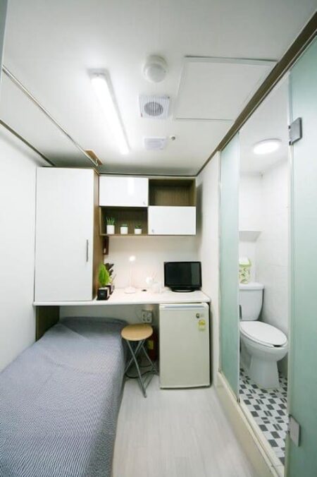小さい部屋の中に必要な家具や設備がよく収まっている効率的な空間。テレビや冷蔵庫、収納棚など、狭い中にも生活に必要なものがきちんと配置されている。白と灰色を基調とした清潔感のある内装も印象的です。