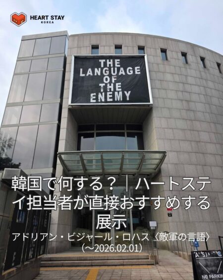アートソンジェセンター入口前で、〈The Language of the Enemy〉展のリストバンド型チケットを手に持つ様子。建物正面には展示タイトルの大型バナーが掲げられている。