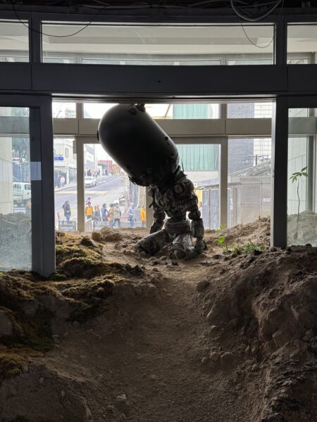 土に覆われた床の上に膝をつく人型彫刻。機械的な身体と頭部を持つ存在が、建物の外の都市風景と対峙している。