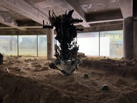 地下空間に設置された巨大な彫刻作品。土や廃材に囲まれ、天井から吊るされた構造物が不安定な生態系を形成している。 