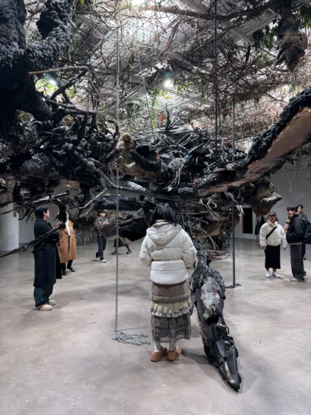 観客が巨大な有機的彫刻の下に立ち、見上げている様子。枝や鎖、廃材が絡み合い、生き物のような存在感を放つ。