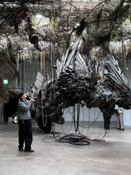 金属や植物、配線が複雑に絡み合った大型彫刻作品。鑑賞者が近くで観察し、没入的な展示空間を体験している。