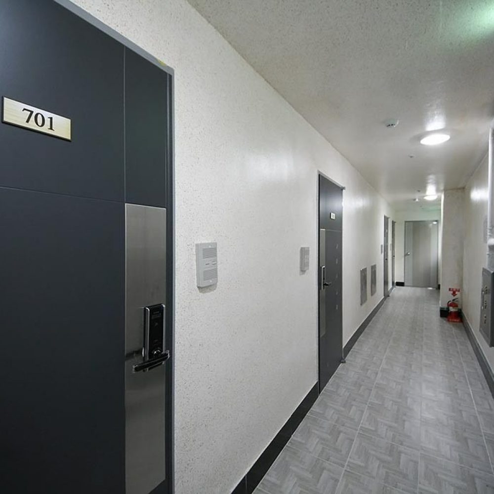 廊下の内部が映った画像です。左側には「701」と表示された暗い色のドアがあり、その隣には別のドアがあります。壁は淡い色合いで、床はタイル状になっています。廊下の奥には消火器が見え、壁に電気設備の箱が取り付けられています。全体的に広々として明るい雰囲気です。