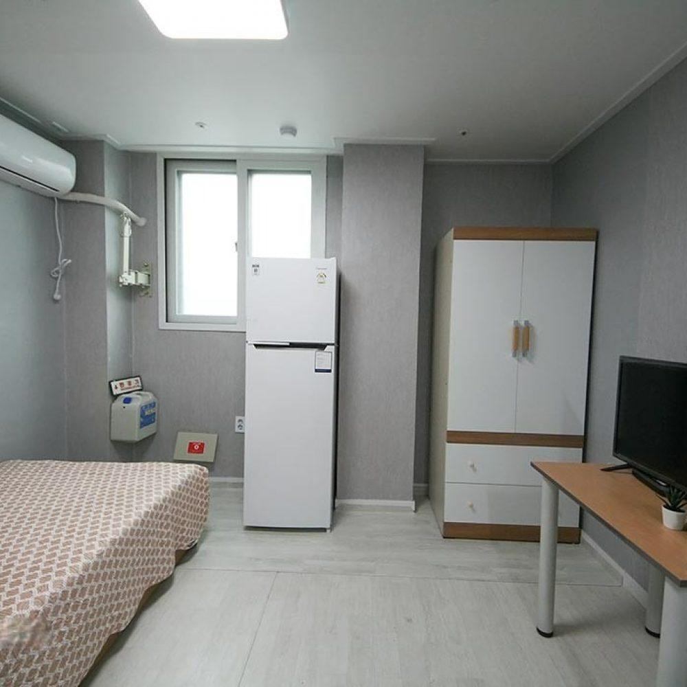 部屋の内部が映った画像です。ベージュの模様が施された布団の掛かったシングルベッドがあり、隣には白い冷蔵庫があります。壁にはエアコンが取り付けられており、窓からは光が差し込んでいます。右側には収納キャビネットがあり、向かいにはテーブルとテレビが設置されています。全体的に明るく、シンプルで快適な雰囲気です。