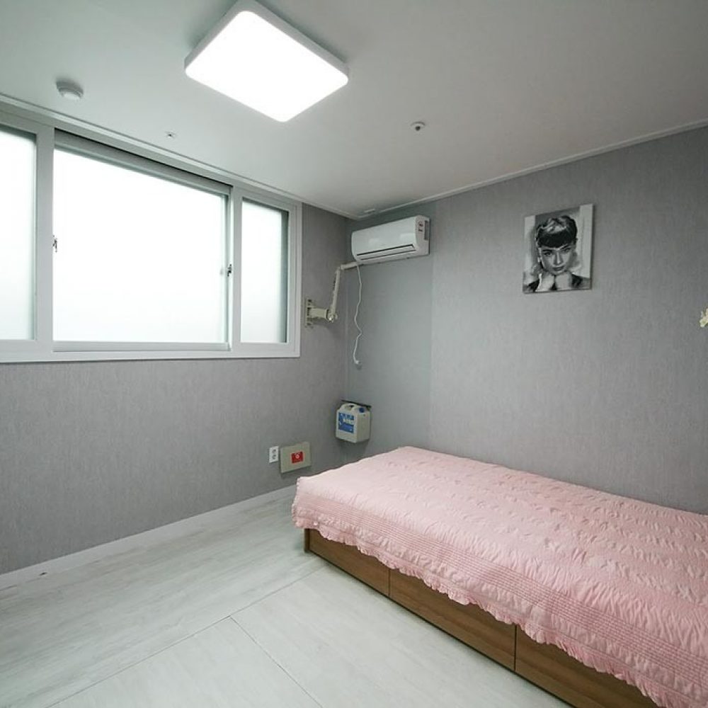 部屋の内部が映った画像です。ピンクの布団が掛けられたシングルベッドがあり、壁には女性のポートレートが飾られています。窓は2つあり、光が差し込む明るい空間です。壁にはエアコンとインターホンも見え、全体的にシンプルで快適な雰囲気です。