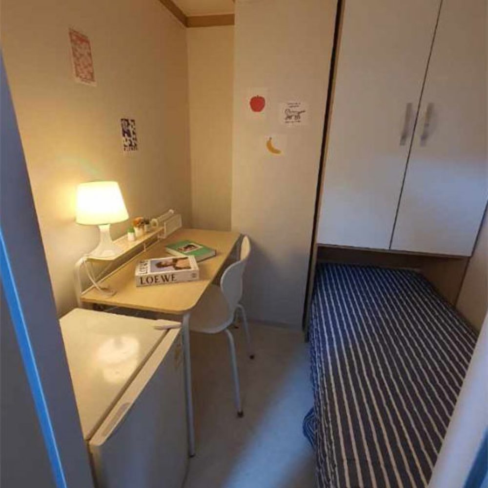 部屋の一角が映った画像です。左側には小さな机と椅子があり、デスクランプが点灯しています。机の上には本や文房具が置かれています。右側にはストライプ模様のシーツがかけられたシングルベッドがあり、その隣に小さな冷蔵庫があります。壁にはいくつかの飾りが貼られており、全体的に温かみのある居心地の良い雰囲気です。