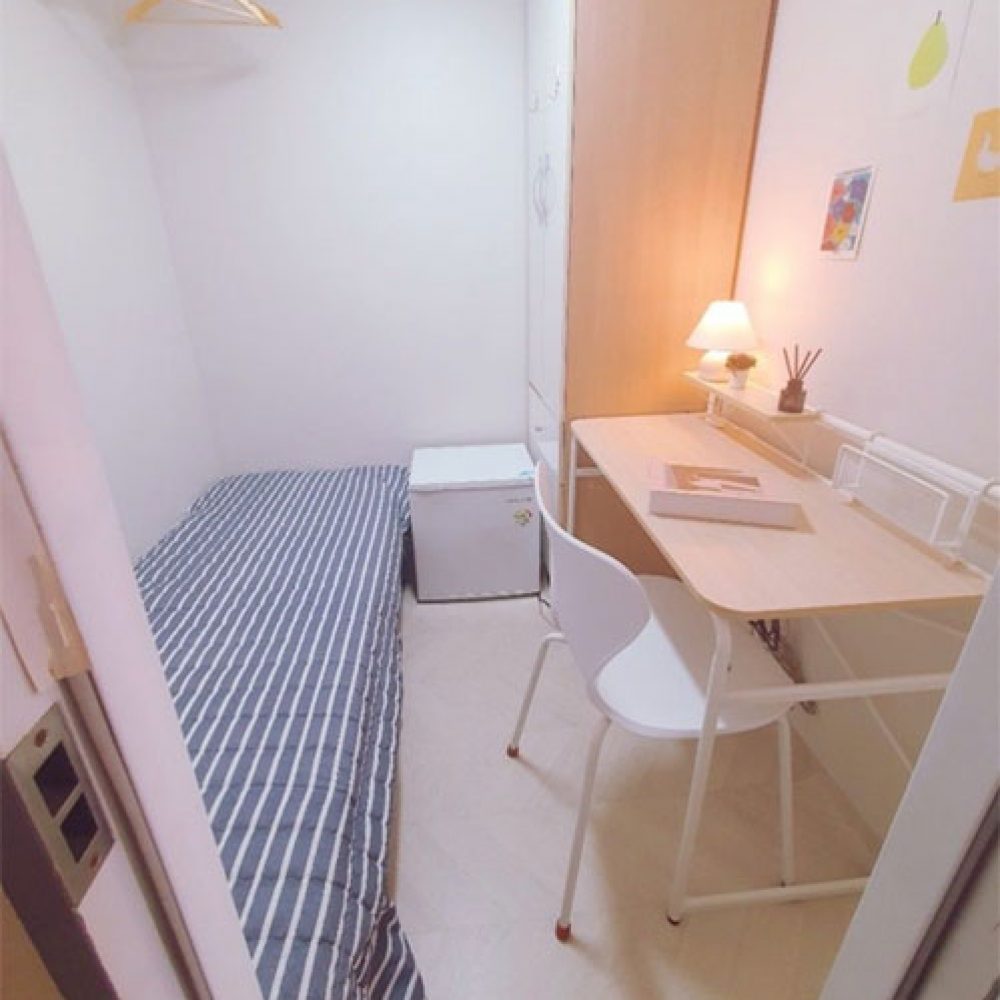 部屋の内部が映った画像です。左側にはストライプ模様のシーツがかけられたシングルベッドがあり、その隣に小さな冷蔵庫があります。右側にはシンプルなデスクと椅子があり、デスクの上にはデスクランプ、いくつかの本、植木鉢があります。また、壁にはカラフルなポストカードが貼られています。全体的に明るく、居心地の良い雰囲気が感じられます。