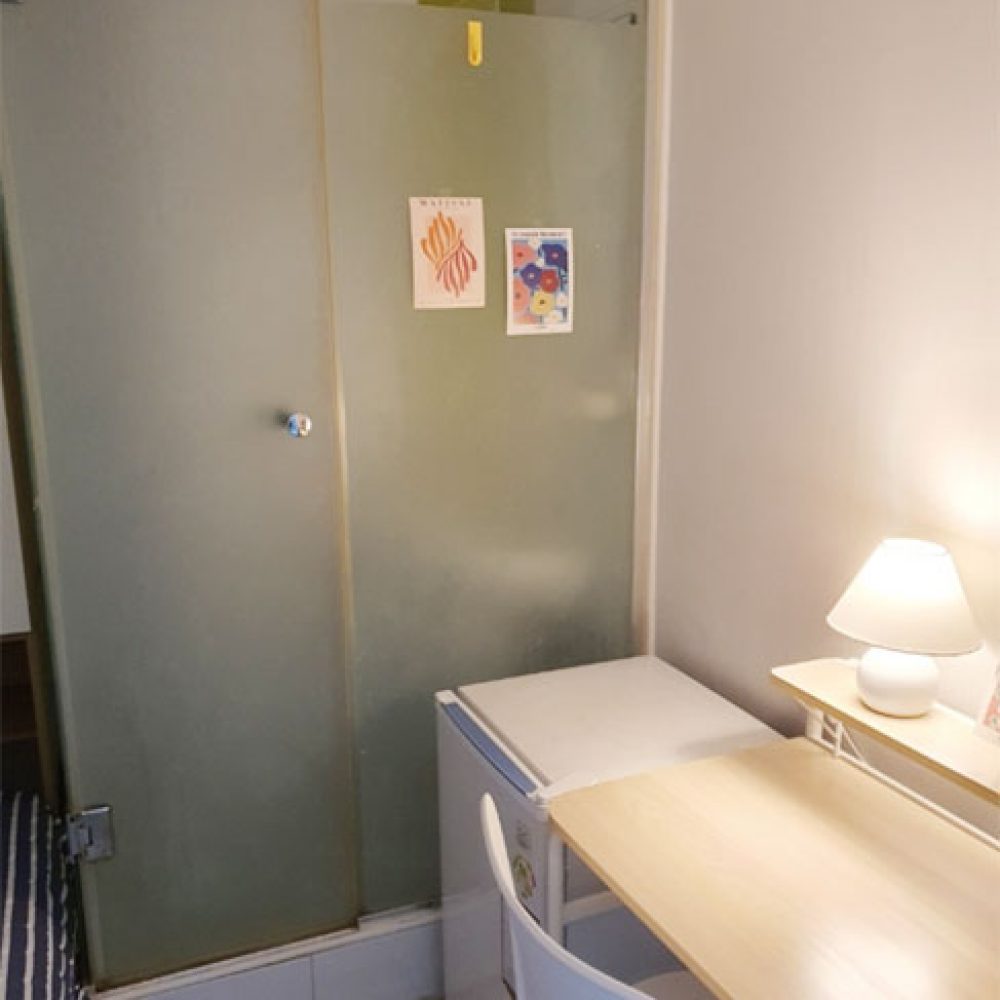 部屋の一角が映った画像です。透明なガラスのシャワールームがあり、その横には小さな冷蔵庫とデスクがあります。デスクの上にはデスクランプがあり、周囲は温かみのある雰囲気です。壁にはいくつかのポストカードが飾られており、全体的にシンプルで居心地の良い印象です。