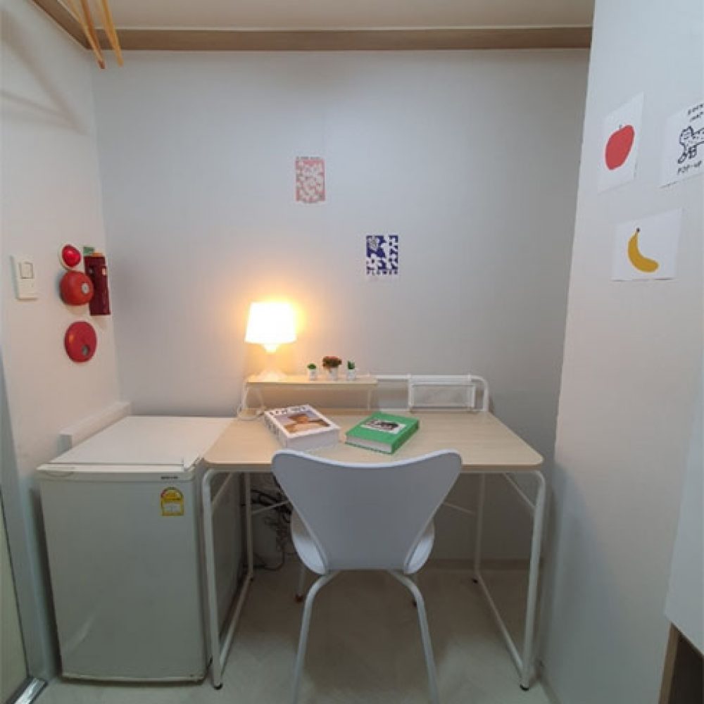 部屋の一角が映った画像です。左側には小さな冷蔵庫があり、その隣にシンプルなデスクがあります。デスクの上にはデスクランプといくつかの本が置かれています。白い椅子がデスクの前にあり、壁にはいくつかのカラフルなポストカードが貼られています。全体的に明るくて居心地の良い雰囲気です。