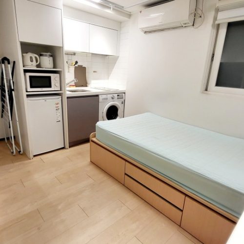 画像は白い内装のキッチンと洗濯機のある部屋を示しています。中央には白と木目調の長いベッドが置かれており、部屋全体が明るく清潔に見えます。