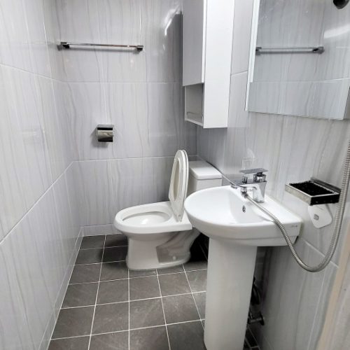 画像は洗面台とトイレがある浴室の写真です。白色のタイルと灰色の床タイルで統一された清潔な空間です。洗面台の上にはタオル掛けや棚が設置されており、機能性も兼ね備えています。このような明るく清潔な浴室は、快適な入浴体験を提供するでしょう。