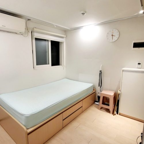 寝室内の写真です。ベッドがあり、ベッドの上に白いマットレスが置かれています。壁には時計がかかっており、部屋には他にも収納棚やエアコンなどの家具が見られます。全体的に明るく清潔な印象の部屋です。