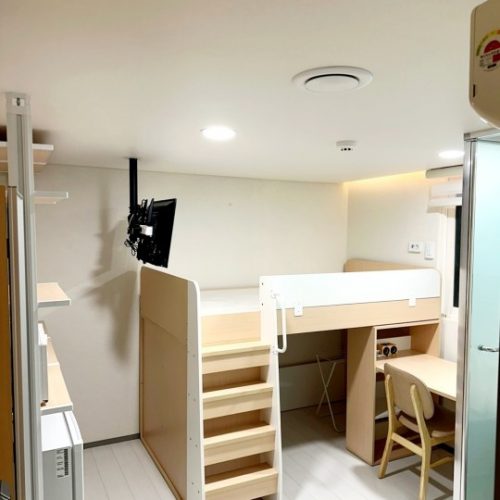 白い壁と天井の明るい一人部屋。壁にはブラケットに取り付けられた小型テレビが設置されています。部屋の中央には、木製で階段付きのロフトベッドが置かれ、下には机と椅子が配置されています。机の上には、電源タップと折りたたみ式の椅子が見えます。部屋の隅には収納棚があり、部屋全体は清潔でシンプルな印象です。