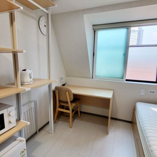 斜めの天井と大きな窓のある、明るく清潔感のあるワンルームの部屋です。窓にはすりガラスのような加工が施されています。部屋の片隅には、木製でシンプルな机と椅子が置かれ、学習スペースとして使えそうです。壁際には、電子レンジと電気ケトルが置かれた白い棚が設置されています。棚の下には、折りたたみ式の物干し台のようなものが見えます。また、右下にはベッドの一部が写っています。全体として、コンパクトながらも機能的な部屋です。