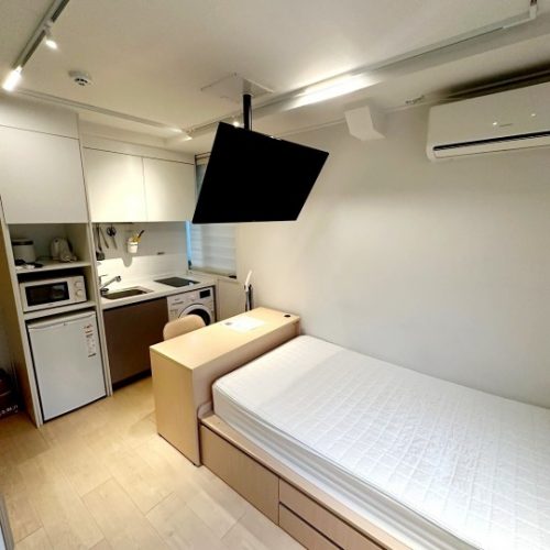 画像には、小さな部屋の内部が写っています。部屋には白い壁、キッチンの設備、ベッド、そして天井に設置された黒いデバイスが見えます。部屋は清潔で整理されており、居心地の良い雰囲気を感じさせます。