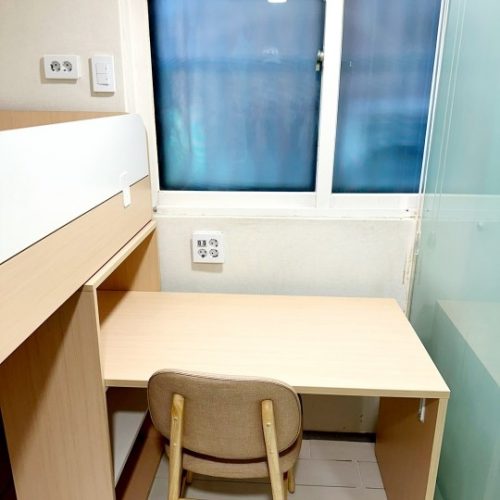 明るい色の木製の学習机と、ベージュの布張り椅子が窓際に置かれています。机はコンパクトで、壁にはコンセントとライトスイッチが複数取り付けられています。机の後ろには、白いヘッドボードの付いたベッドの一部が見えます。窓の外は青みがかった景色で、ガラス戸が二枚並んでいます。右端にはガラスの仕切りが見えます。全体として、学生寮の部屋の一角のような印象です。