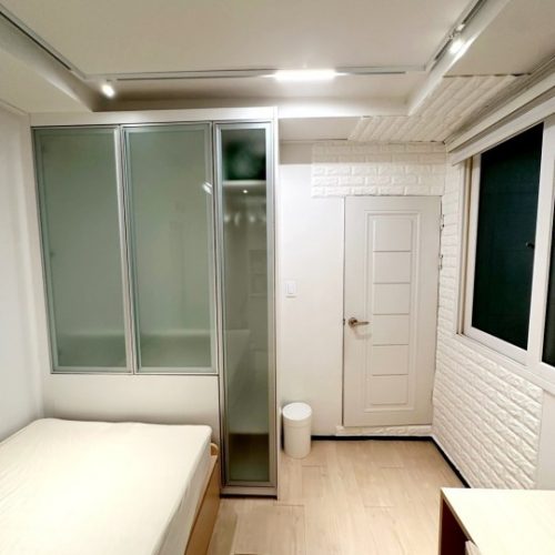 部屋の様子は明るく清潔な感じです。ドア、壁、窓などの内装がシンプルで落ち着いた雰囲気です。部屋の中央には白い寝具が置かれており、収納スペースも確保されているようです。全体的に居心地の良い空間になっていると感じられます。
