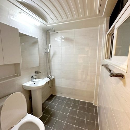 浴室の画像です。明るい白い色調で、トイレと洗面台、シャワーの設備が見えます。天井は格天井になっており、清潔で機能的な印象の空間です。