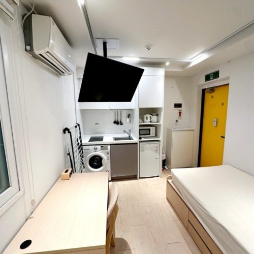 この画像は、マイクロホテルまたはスタジオタイプのアパートメントのインテリアを表しています。部屋は小さいながらも、必要な家電製品やキッチン用品が備えられていて、非常に使い勝手が良さそうです。壁の色は白で明るく、木目調の家具が温かみのある雰囲気を演出しています。ドアの色も鮮やかな黄色で、コンパクトながら居心地の良い空間になっていると思います。