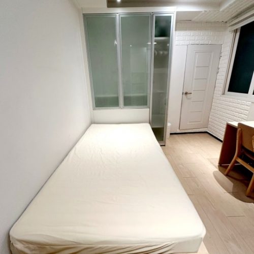 この画像は、白い壁と床の部屋の様子を示しています。部屋には大きな窓があり、白い寝台が置かれています。部屋の奥には白い扉が見えます。全体的に明るく整理された空間です。