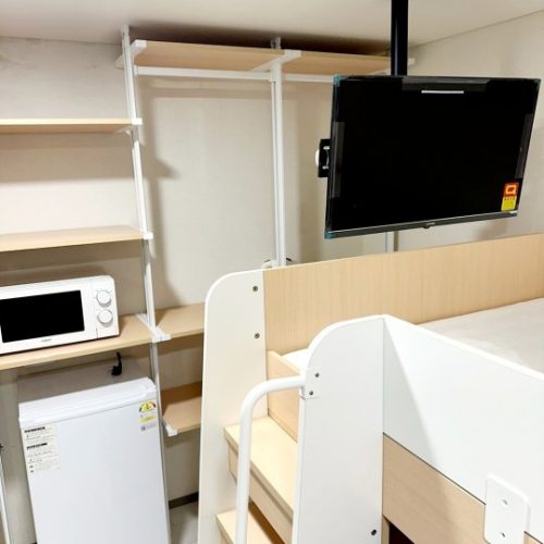 白と木製の家具が配置された部屋の隅を写した写真です。天井から吊り下げられたフラットスクリーンテレビが壁に設置されています。その下に、二段ベッドがあり、上段は木製のフレーム、下段は白く塗装されたフレームです。ベッドの隣には、白い小型冷蔵庫と電子レンジが置かれた白い棚が設置されています。棚の上には、電子レンジと冷蔵庫が置かれています。棚の後ろには、白い金属製のポールと木製の棚からなる収納棚があります。全体的に明るく清潔感のある空間です。
