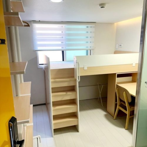 木製のロフトベッドと机、収納棚が配置された、清潔感のある一人部屋です。ベッドには階段状の昇降口があり、机の下には椅子が置かれています。窓には白と淡いブルーのブラインドが取り付けられています。部屋の壁は白で、黄色い収納棚が壁際に設置されています。