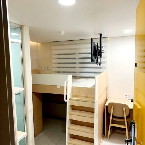ベージュの壁と天井があり、明るい部屋 壁に白と黒のストライプの模様のカーテンがかかっている 部屋の中央には木製の家具が置かれ、引き出しと棚がある 部屋の奥には窓があり、外の景色が見えている 家具の上には装飾品や小物が置かれている 部屋全体が清潔で整頓された雰囲気