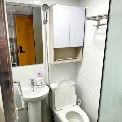 コンパクトなホテルのバスルーム。白い壁とタイルの床、白い洗面台、トイレ、そして壁掛けの棚が清潔感を演出しています。シャワーヘッドは手元で操作できるタイプで、鏡とタオル掛けも設置されています。黄色のドアが写った鏡から、部屋の様子も伺えます。全体的にシンプルで機能的な空間です。