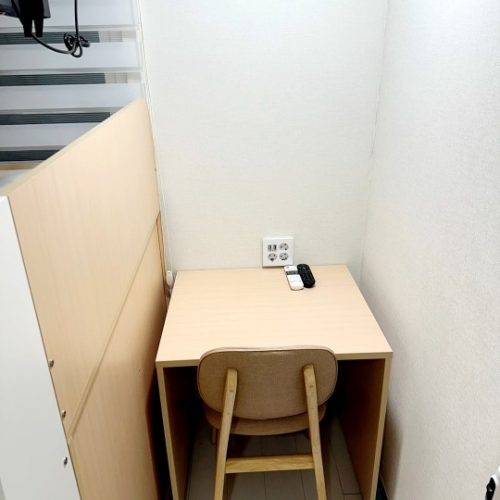 壁に沿って置かれた木の机と、それに合わせた色の椅子。机の上にはコンセントと2つのリモコンが置かれている。壁は白く、シンプルな空間である。