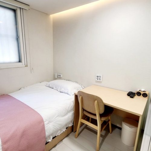 シンプルな一人部屋の画像です。 ベッド、机、椅子が配置されています。ベッドにはピンク色の毛布がかけられています。窓からは自然光が入っています。壁は白く、清潔感のある部屋です。机の上には、電話と小さな収納ボックスが置かれています。全体的にシンプルで落ち着いた雰囲気の部屋です。