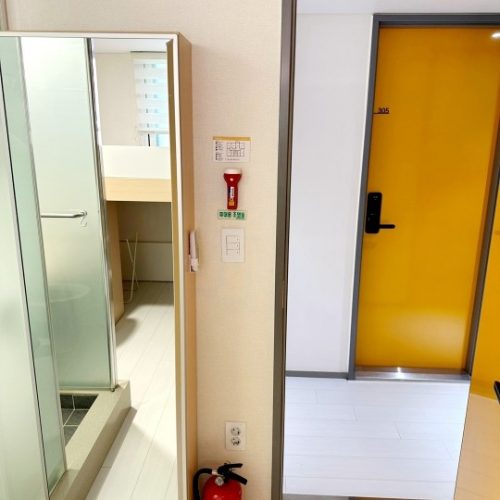 室内の一角を写した写真です。廊下の壁には、黄色い扉（305号室と番号が記されている）と、消火器、懐中電灯、非常口表示が設置されています。壁の反対側には、ガラスの仕切りで区切られたシャワー室と洗面所のある部屋の一部が見えます。部屋の床は白っぽい木目調です。