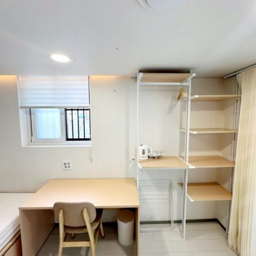 白を基調とした清潔感のある部屋。窓にはブラインドが設置され、机と椅子が置かれています。壁際には、棚とハンガーラックが一体となった収納家具があり、電気ケトルなどが置かれています。全体的にシンプルで機能的な空間です。