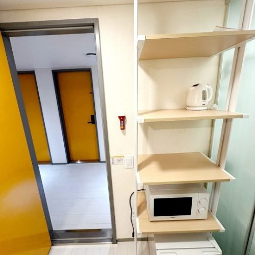 白色のシンプルな棚には、電子レンジと白い電気ケトルが置かれています。棚は部屋の壁際に設置され、棚の後ろには黄色のドアが並んだ廊下が見えます。部屋は清潔で整然としています。