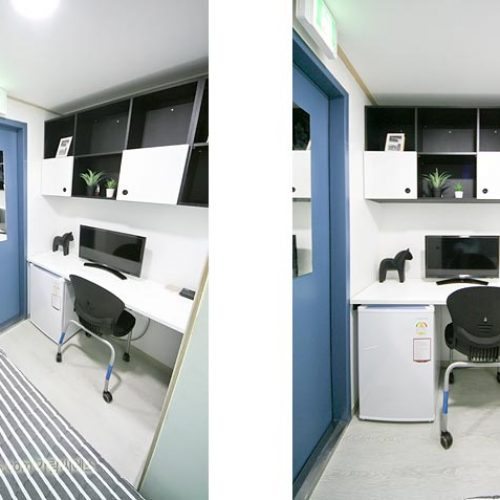 画像は小さな部屋の内部を示しています。部屋には白と黒の色調が使われています。壁に棚が設置され、テレビやその他の電子機器が置かれています。部屋の中央には机と椅子が置かれ、作業スペースとして使われているようです。全体的に整理された印象を受けます。