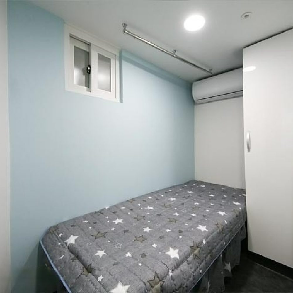 この画像は、明るい青色の壁を持つ小さな部屋の内部を示しています。部屋の左側には、星柄のブランケットがかけられたベッドがあります。右側には収納スペースがあり、上部にはエアコンが設置されています。また、左上には小窓があり、部屋は明るい照明で照らされています。