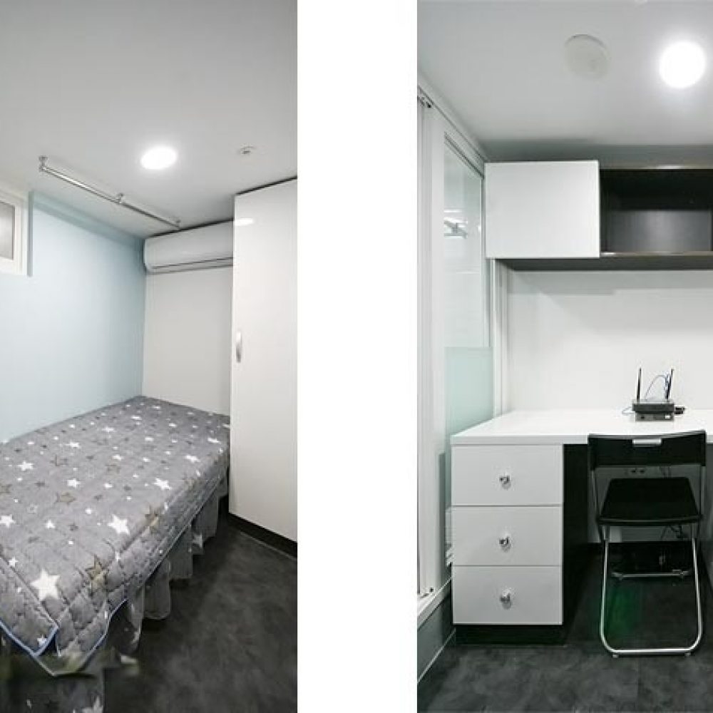 この画像は、左側にベッドが置かれた部屋の部分と、右側にデスクと冷蔵庫がある作業スペースの部分で構成されています。部屋は明るい色調で、シンプルで機能的なデザインが特徴です。ベッドには星柄のブランケットがかけられています。デスクの上にはモニターや文房具が置かれており、下には引き出しと冷蔵庫があります。